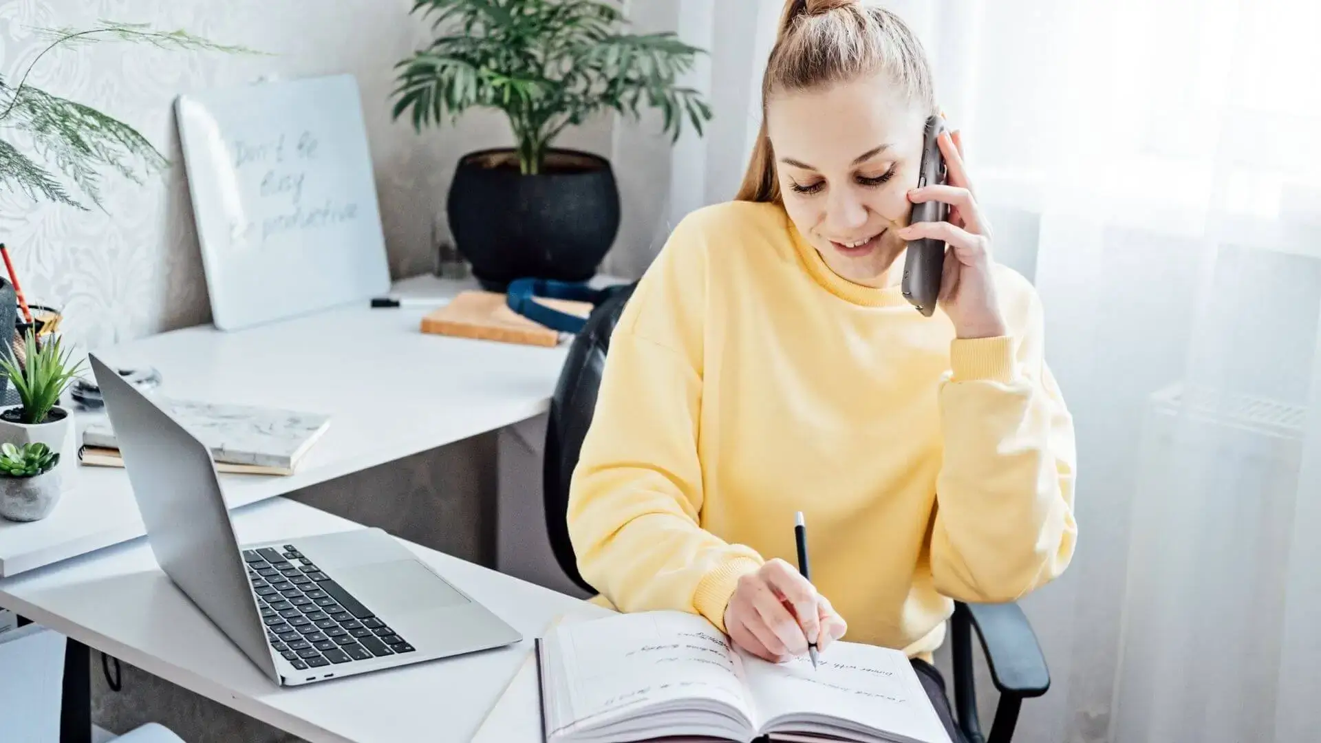 flexibilite-adaptabilite-placements-financiers-lyon Jeune femme au téléphone prenant des notes à son bureau, avec un ordinateur portable, pour illustrer la flexibilité et l’adaptabilité dans le suivi des placements financiers.