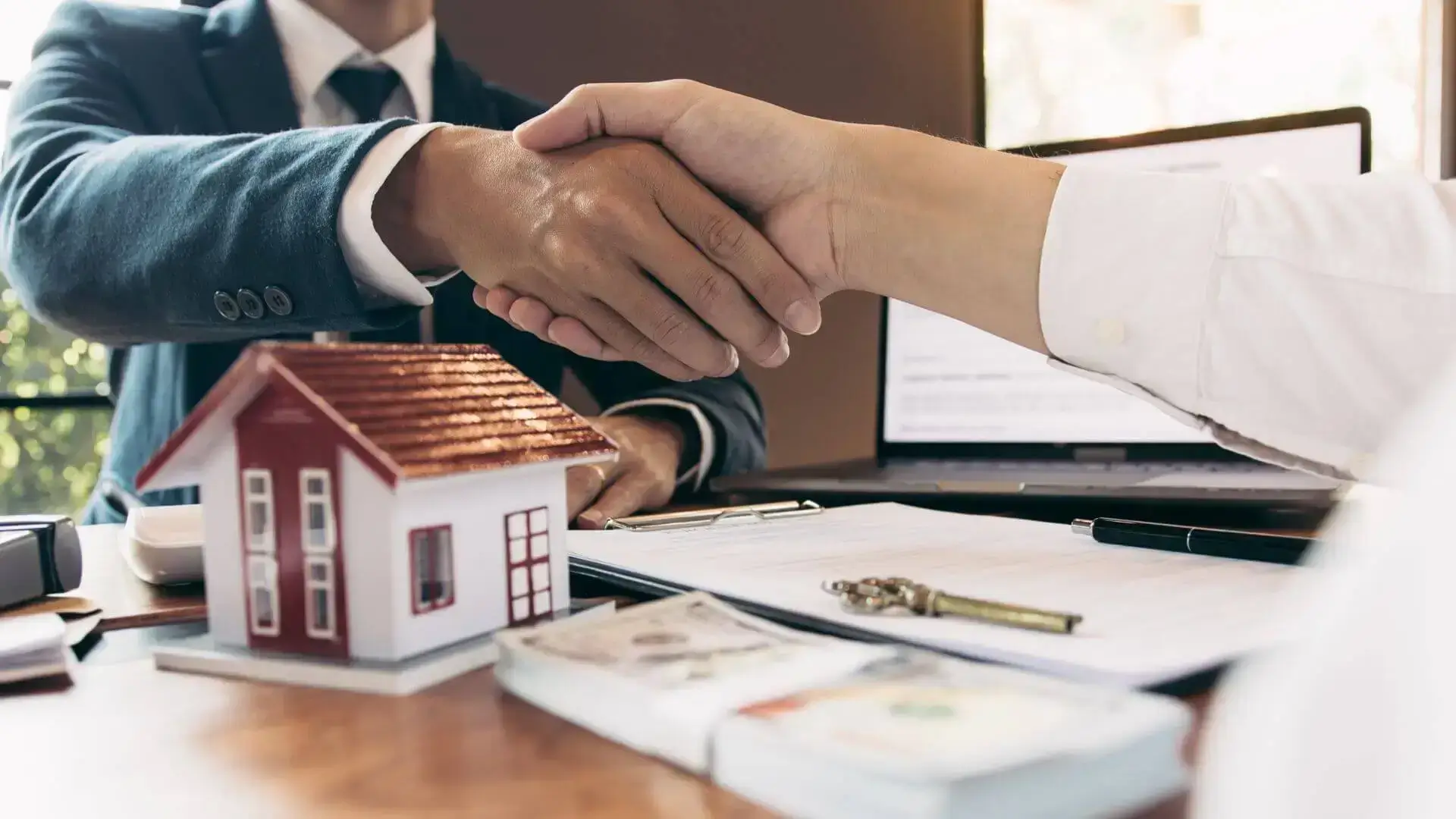 Deux personnes se serrent la main devant une maison miniature, des clés et des documents, pour illustrer la gestion des transactions immobilières.