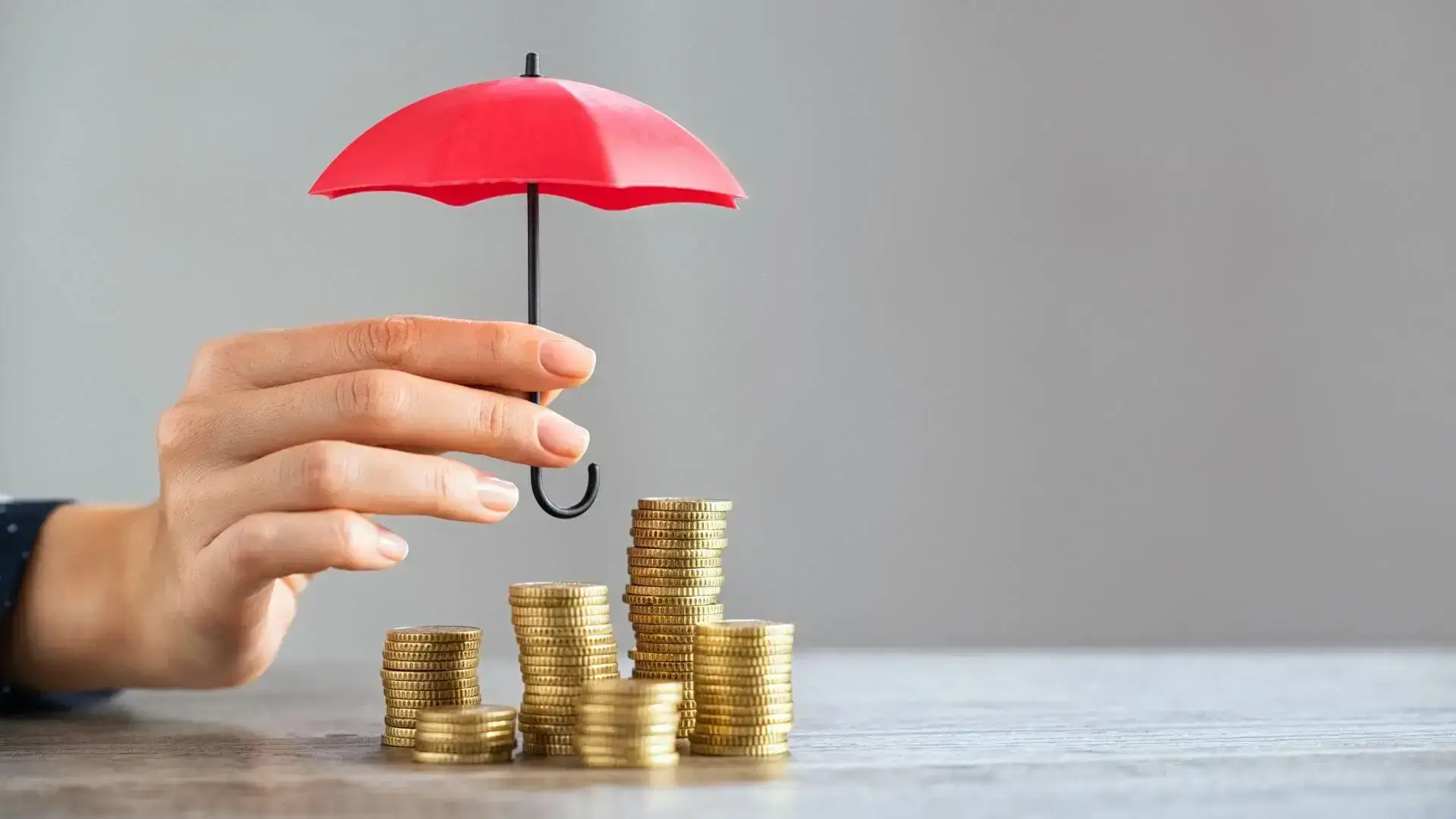 Main tenant un parapluie rouge au-dessus de piles de pièces, pour illustrer la sécurité du patrimoine et la protection financière.