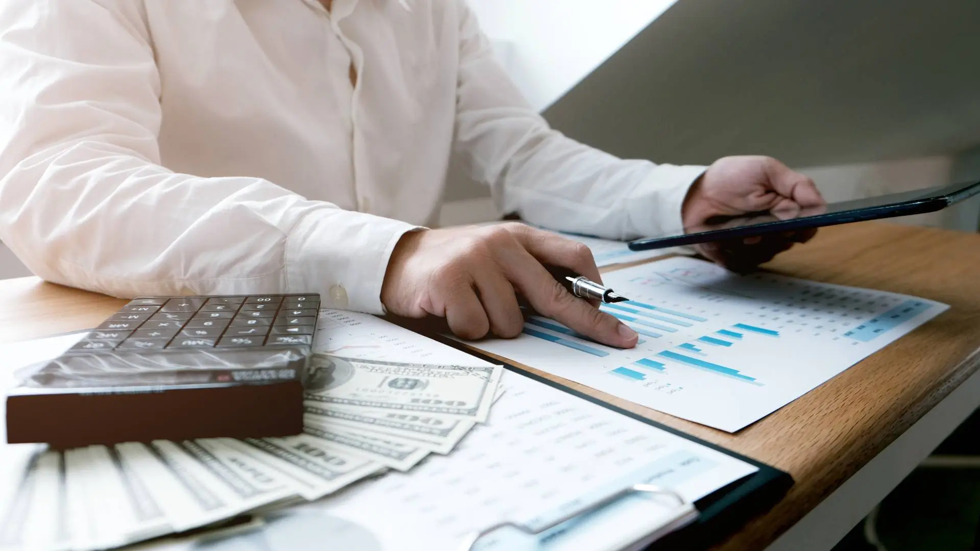 Personne analysant des graphiques financiers avec un stylo, une tablette, une calculatrice et des billets, pour illustrer la création de revenus passifs grâce à l’investissement immobilier.